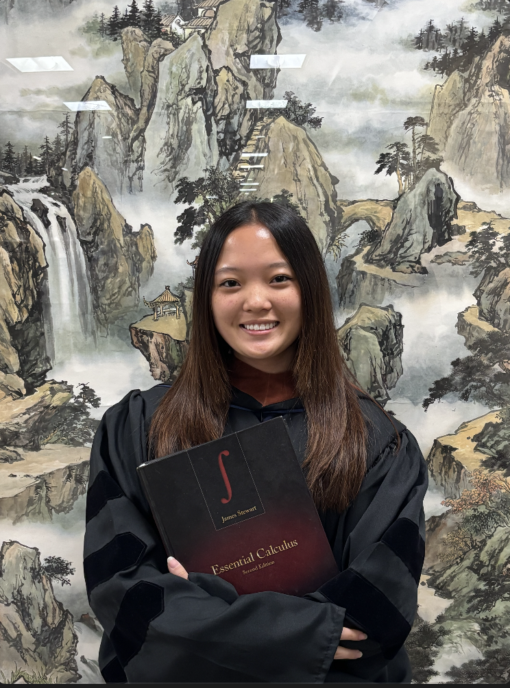 Professor Ainsworth stands in black before a painting and holds a book.